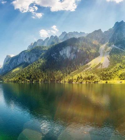 Paisaje de un lago en el que se reflejan las montañas que tiene junto a él con bosque y praderas verdes. Todo ello está iluminado con rayos de sol